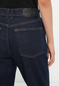Temno denim hlače z gladko teksturo, povišanim pasom in vidnim šivanjem. Imajo označen črn usnjen pečat na hrbtu.