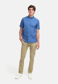 Kortermet lyseblå skjorte med krage, kombinert med beige bukser og hvite joggesko, stående mot en ensfarget bakgrunn.