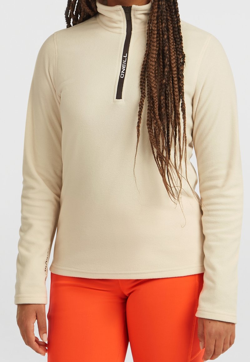 Vrouw draagt een beige fleece-trui met een lange mouw en een kwartritssluiting met zwarte rits, en felle oranje legging, staand tegen een effen achtergrond.