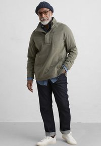 Chaqueta pullover verde oliva con media cremallera, detalles de bolsillo frontal, combinada con una camisa de denim azul clara, jeans oscuros y zapatillas blancas.