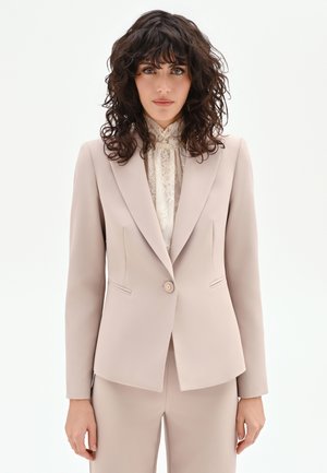 Femme aux cheveux bouclés et foncés portant une veste de costume beige sur un chemisier blanc en dentelle à col montant, debout devant un fond blanc uni.