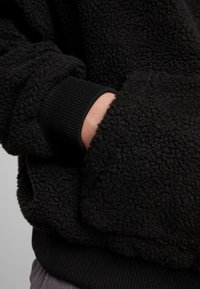 Sweatshirt en polaire noir texturée avec un extérieur doux, des poignets côtelés et une main dans une poche latérale. Le matériau semble chaud et confortable.