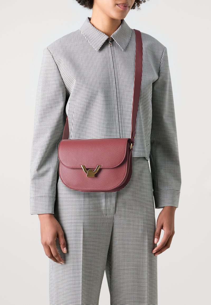 Maroon leather crossbody bag with a textured finish; features a gold-tone clasp and adjustable strap. Worn over a checkered garment.