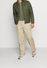 Giacca a vento verde con design a righe orizzontali, chiusura con zip, abbinata a un polo verde scuro, chinos beige e sneaker chiari.