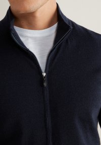 Granatowy sweter z zamkiem błyskawicznym, wykonany z miękkiego materiału, z wysokim kołnierzem i kontrastowym zamkiem z metalowym uchwytem. Nosi się go na białej koszuli.