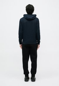 Tengerészkék kapucnis pulóver nagy kapucnival és bordázott szegéllyel, fekete melegítőnadrággal párosítva. Bal ujján logónyomattal. Fekete lábbelivel.