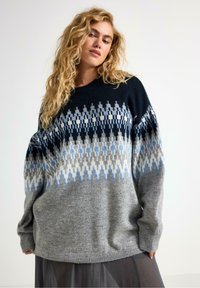 Donna con lunghi capelli ricci che indossa un maglione in maglia a vestibilità ampia con un motivo geometrico blu, grigio, nero e bianco sopra una gonna scura.