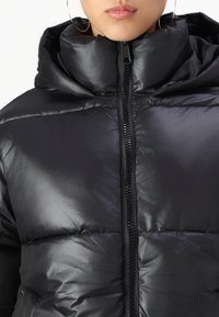 Čierna puffer bunda s vysokým golierom, lesklý materiál a viditeľné švy. Predný zips a nadmerná kapucňa pre zvýšené teplo.