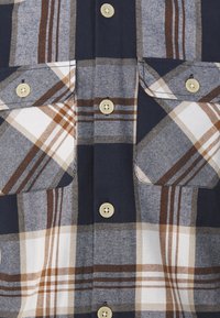 Camisa de cuadros azul y blanca con acentos marrones, que presenta grandes bolsillos, botones crema y una tela suave y texturizada.