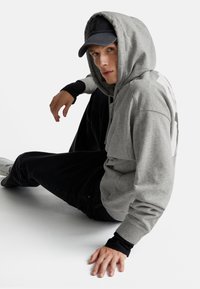 Un giovane in felpa grigia e cappellino nero è seduto sul pavimento con un ginocchio sollevato, mentre guarda leggermente di lato.