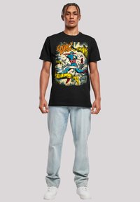 Svart bomull t-skjorte med et fargerikt tegneserie-trykk på forsiden. Modellen har på seg lyseblå jeans og hvite joggesko.