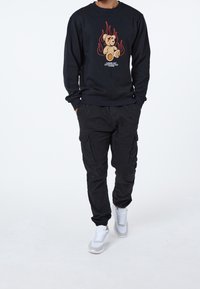 Suéter azul marino con un gráfico de un oso rodeado de llamas, combinado con pantalones cargo negros y zapatillas blancas. Atuendo casual y holgado.