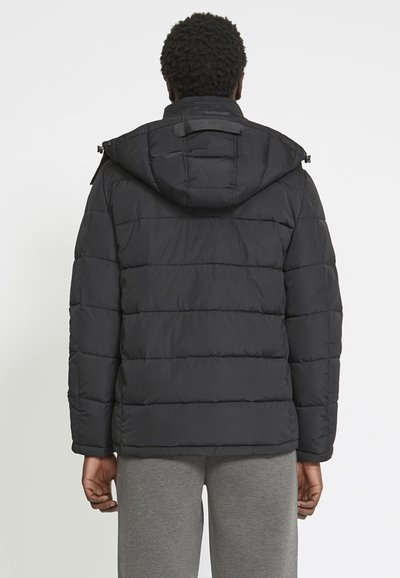 Giacca a vento nera con cappuccio, caratterizzata da pannelli trapuntati orizzontali, zip frontale e una toppa vicino al colletto. Indossata con pantaloni grigi.