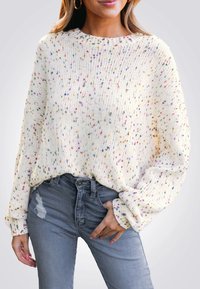 Chunky gebreide trui in gebroken wit met meerkleurige spikkels, voorzien van een geribde halslijn en brede mouwen, gecombineerd met versleten grijze jeans.