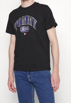 Personne portant un t-shirt noir Tommy Jeans avec un logo bleu et beige et un jean bleu, debout devant un fond blanc uni.