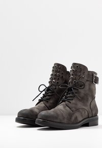 Bottines en cuir suédé gris ornées de clous ronds noirs sur le col, avec lacets à l'avant et un petit talon bloc. Matière texturée avec des détails de coutures.