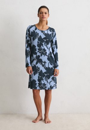 Frau, die barfuß steht und ein knie-langes hellblaues Kleid mit großen schwarzen Blumenmustern trägt, vor einer schlichten weißen Wand.