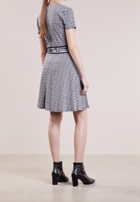 Zwart-wit houndstooth jurk met korte mouwen, een uitlopende rok en een zwarte, gevlekte riem. G gedragen met zwarte enkellaarsjes. Gladde textuur.