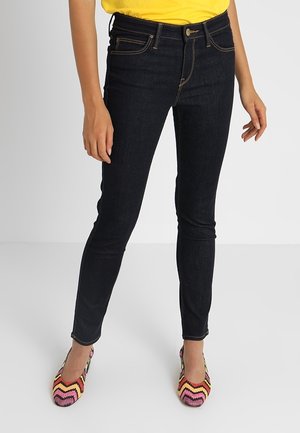 Jeans oscuros delgados de color azul con una textura suave, que cuentan con cinco bolsillos, un cierre de botón y detalles de costura, combinados con unos zapatos planos multicolores.