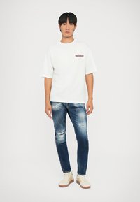 T-shirt bianca a maniche corte con il testo rosso "DSQUARED2" sul petto, abbinata a jeans in denim blu scuro sfilacciati e sneakers beige.