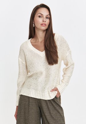 Sweter w kolorze kremowym z dekoltem w serek, z teksturowanym wzorem, z prążkowanymi mankietami i dołem; noszony na wzorzyste spodnie o wysokim stanie.