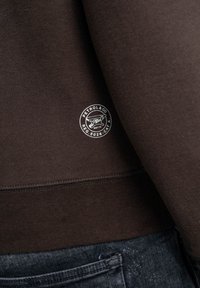 Hnědá mikina z měkké látky, s kulatou bílou nášivkou loga na spodní straně a žebrovaným lemem.