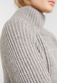 Strickpullover mit Rollkragen in sanftem Grau, ausgestattet mit gerippter Textur und dezenten Kabeldetails an der Schulter für zusätzlichen visuellen Reiz.