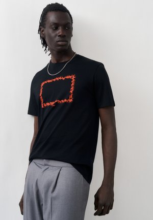 Νέος άνδρας που φοράει μαύρο μπλουζάκι με πορτοκαλί ορθογώνιο σχέδιο μυρμηγκιού και ανοιχτό γκρι πλισέ παντελόνια, κοιτάζοντας προς τα πλάγια.