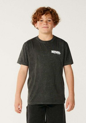Niño con cabello rizado rojo y pecas que lleva una camiseta negra lavada oscura y pantalones cortos a juego, de pie frente a un fondo liso.