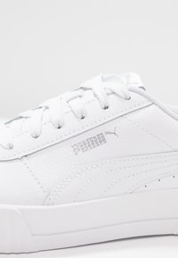 Vit Puma-sneaker med texturerat läder, vita snören och silverfärgad Puma-logga på sidan, visad mot en enfärgad vit bakgrund.