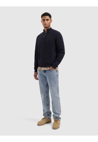 Marineblauwe gebreide trui met een kwart ritssluiting, gecombineerd met een beige gelaagde shirt, versleten lichtblauwe jeans en tan geknoopte schoenen.