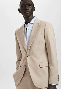 Jasno beżowy garnitur wykonany z tkaniny o gładkiej fakturze, z klapami w szpic, jedno zapięcie z przodu oraz jasnoniebieską koszulą pod spodem.