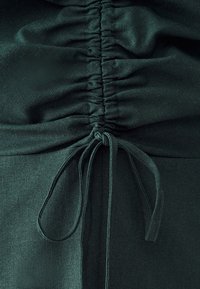 Dark green fabric blouse featuring gathered detailing at the waist and a tie closure for adjustment. Smooth texture with a slight sheen.