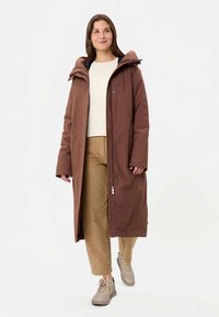 Brūna gara pūkaina jaka ar kapuci, aizdares piespraudes, sānu kabatas, valkāta virs gaiša topa un bēšām biksēm, kopā ar bēšām sporta kurpēm.