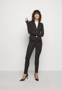 Blazer nero aderente con bottoni, abbinato a pantaloni slim-fit coordinati. Indossato con un top bianco e tacchi a spillo neri. Texture liscia.