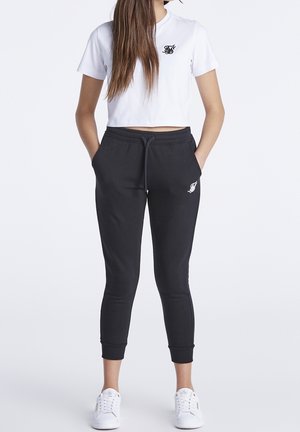 Femme portant un t-shirt court blanc, un pantalon de jogging noir avec cordon de serrage et des baskets blanches, debout les mains dans les poches sur un fond uni.