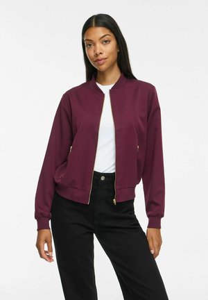 Femme portant une veste bordeaux zippée par-dessus un haut blanc et un pantalon noir, debout dans une posture détendue et confiante sur un fond uni.
