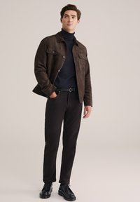 Jonge man met een donkerbruine suède jas, een marineblauwe coltrui, zwarte broek, een zwarte geweven riem en zwarte net shoes, staand op een beige achtergrond.
