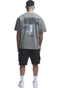 Homme portant un t-shirt gris oversized avec le texte « INTROSPECT Yourself » et un visuel de téléphone vintage, assorti à un short cargo noir et des baskets blanches.