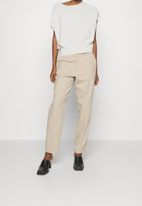 Béžové kalhoty na míru v kombinaci s bílým topem s krátkým rukávem. Modelka má na sobě černé lodičky na podpatku, které představují čistý, minimalistický design.