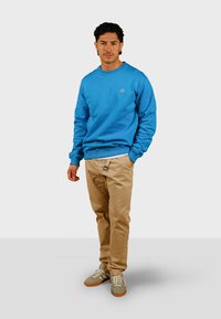 Modrá bavlnená mikina s rebrovými manžetami a lemom, kombinovaná s béžovými chinos nohavicami a šedými teniskami. Jednoduchý logo detail na mikine.