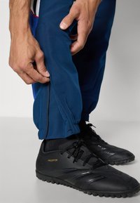 Calças de atletismo navy com aberturas em zip na parte inferior das pernas e punhos elásticos, combinadas com sapatilhas de futebol pretas apresentando o logótipo dourado "Predator".