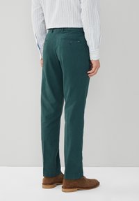 Pantalon en coton vert à coupe droite. Il comporte une poche arrière avec fermeture à bouton et est porté avec des chaussures en daim marron.