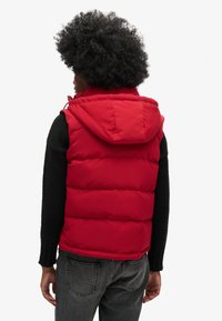 Chaleco acolchado rojo con capucha, que presenta una textura suave y costuras horizontales, usado sobre una camiseta negra de manga larga y jeans grises.