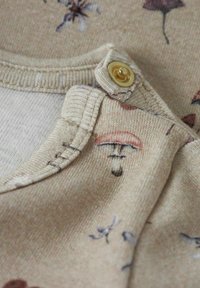 Beige tyg med blommönster, med rundad halsringning och en guldknäppning nära axeln. Texturerad sömdetail synlig.