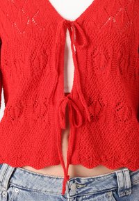 Cardigã vermelho de malha com bainha recortada, frente aberta e fecho com laço. Apresenta um padrão de rendas e tecido texturizado. Usado sobre uma blusa clara.