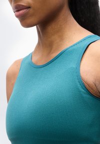 Κοντινό πλάνο ενός ατόμου που φοράει μια τυρκουάζ ριμπ μπλούζα χωρίς μανίκια, εμφανίζοντας τον λαιμό, τον ώμο και μέρος του άνω στήθους σε λευκό φόντο.