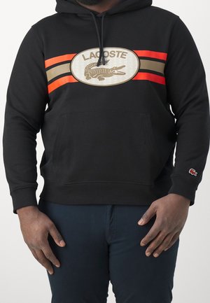 Sweat à capuche noir avec des rayures rouges et beiges et un grand logo Lacoste représentant un crocodile sur la poitrine, porté par une personne à la peau foncée.