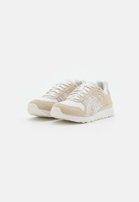 Beige suede and mesh sneakers with white accents, round toe, smooth sole, and prominent brand logo on the side. Laced design adds detail.