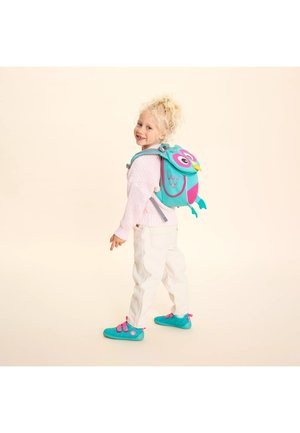 Junges Kind lächelt, trägt einen rosa Pullover, weiße Hose, türkisfarbene Schuhe und einen bunten, eulenförmigen Rucksack, steht vor einem hellen Hintergrund.